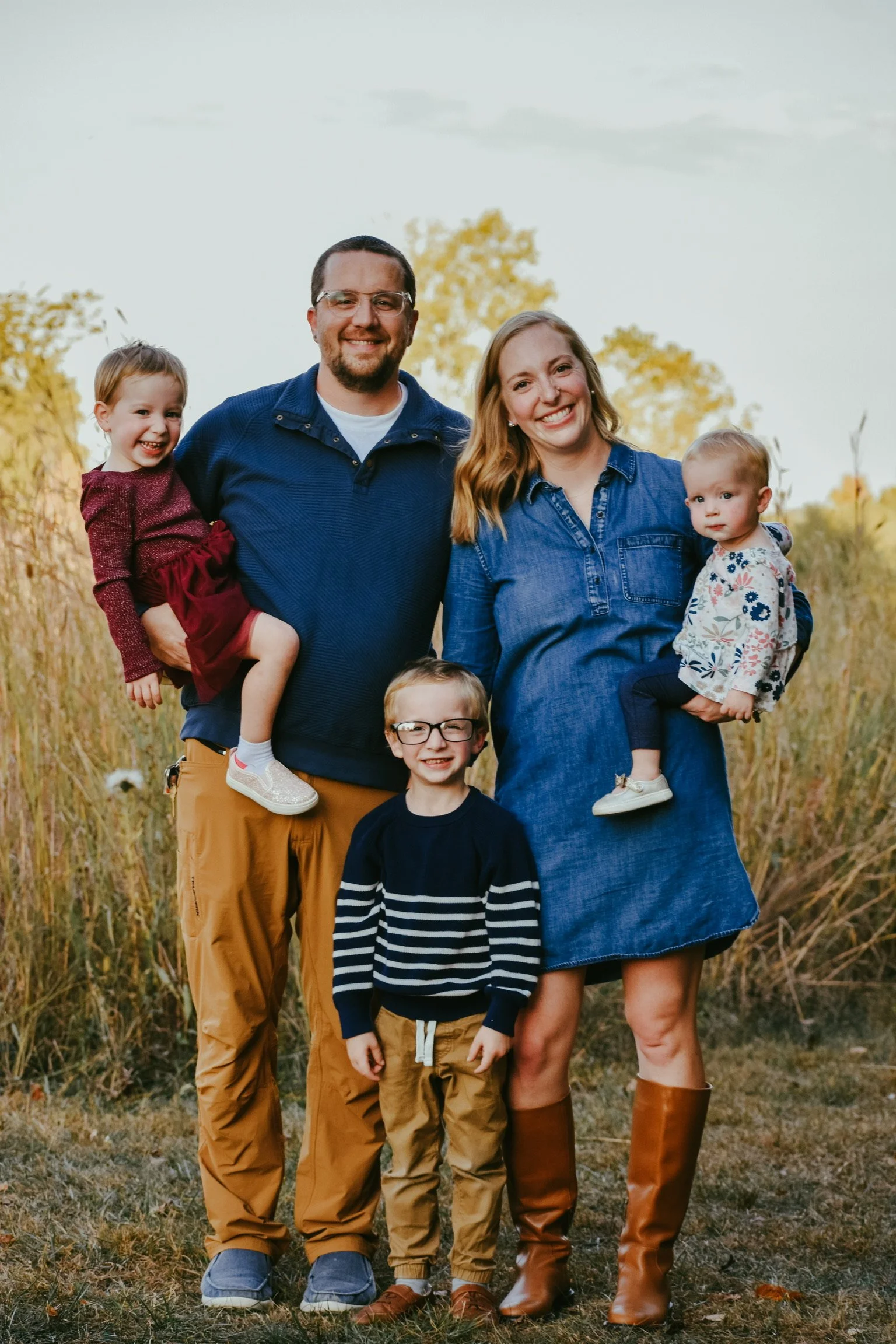 Family of five standing outdoors in a field with tall grass and trees in the background, smiling at the camera. The family includes a man, woman, and three children, each dressed casually and warmly.