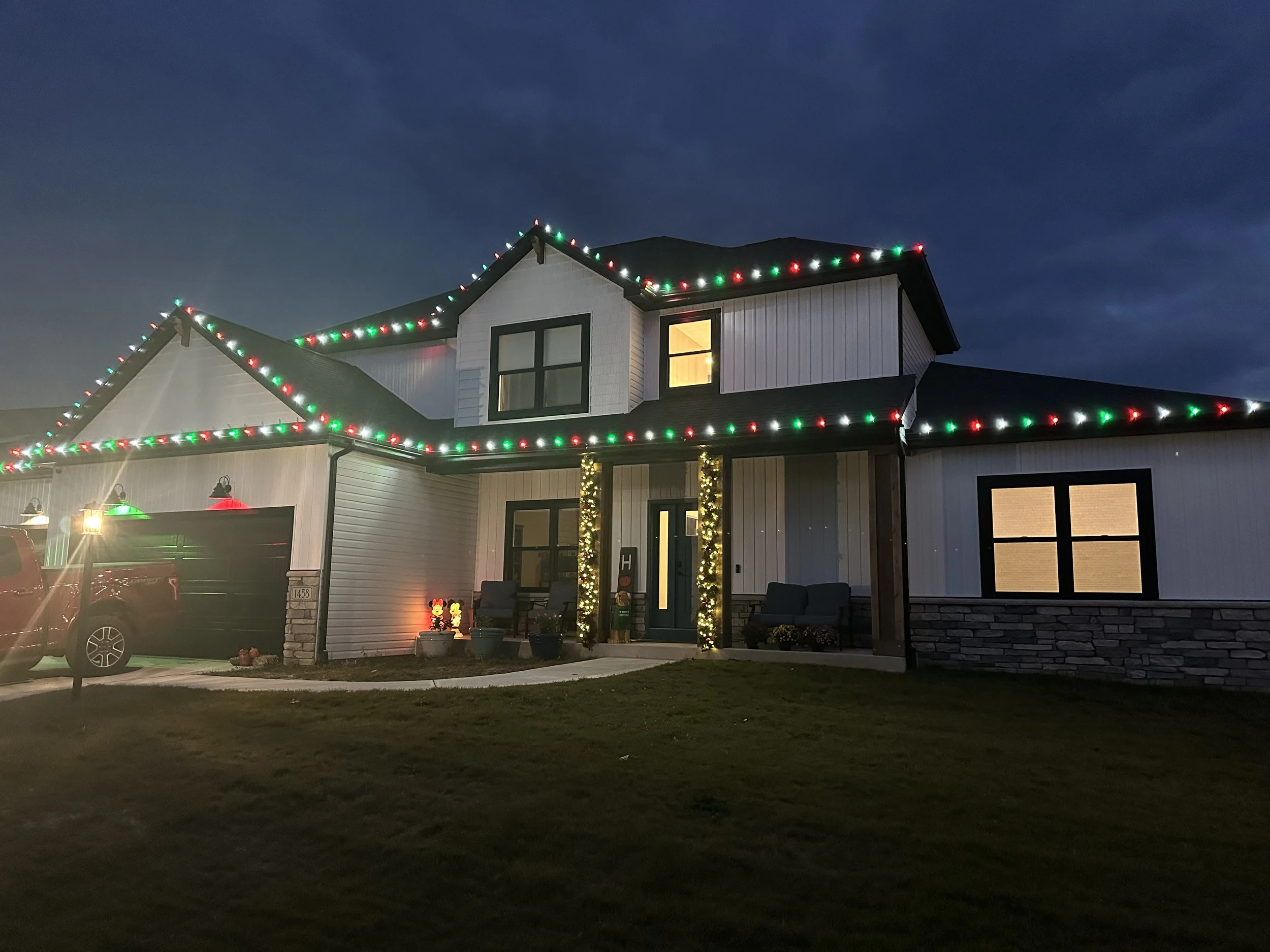 A house decorated with Christmas lights, with a dark blue sky in the background. The house has multiple windows, some with interior lighting visible, and string lights along the roofline in red, green, and white. There is a small front porch with chairs and holiday decorations, and the lawn is partly visible with some outdoor seating.