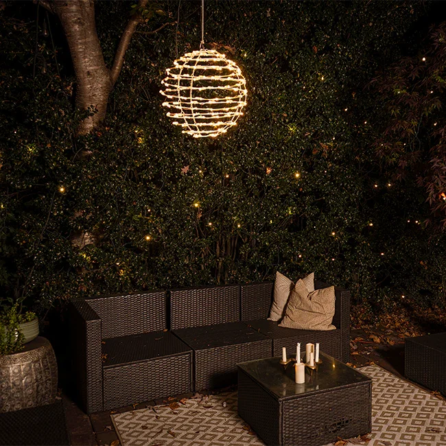 Outdoor patio seating area at night with a black wicker couch, pillows, a black wicker coffee table with candles, a decorative rug, and a glowing spherical lantern hanging from a tree. There are small string lights in the background.