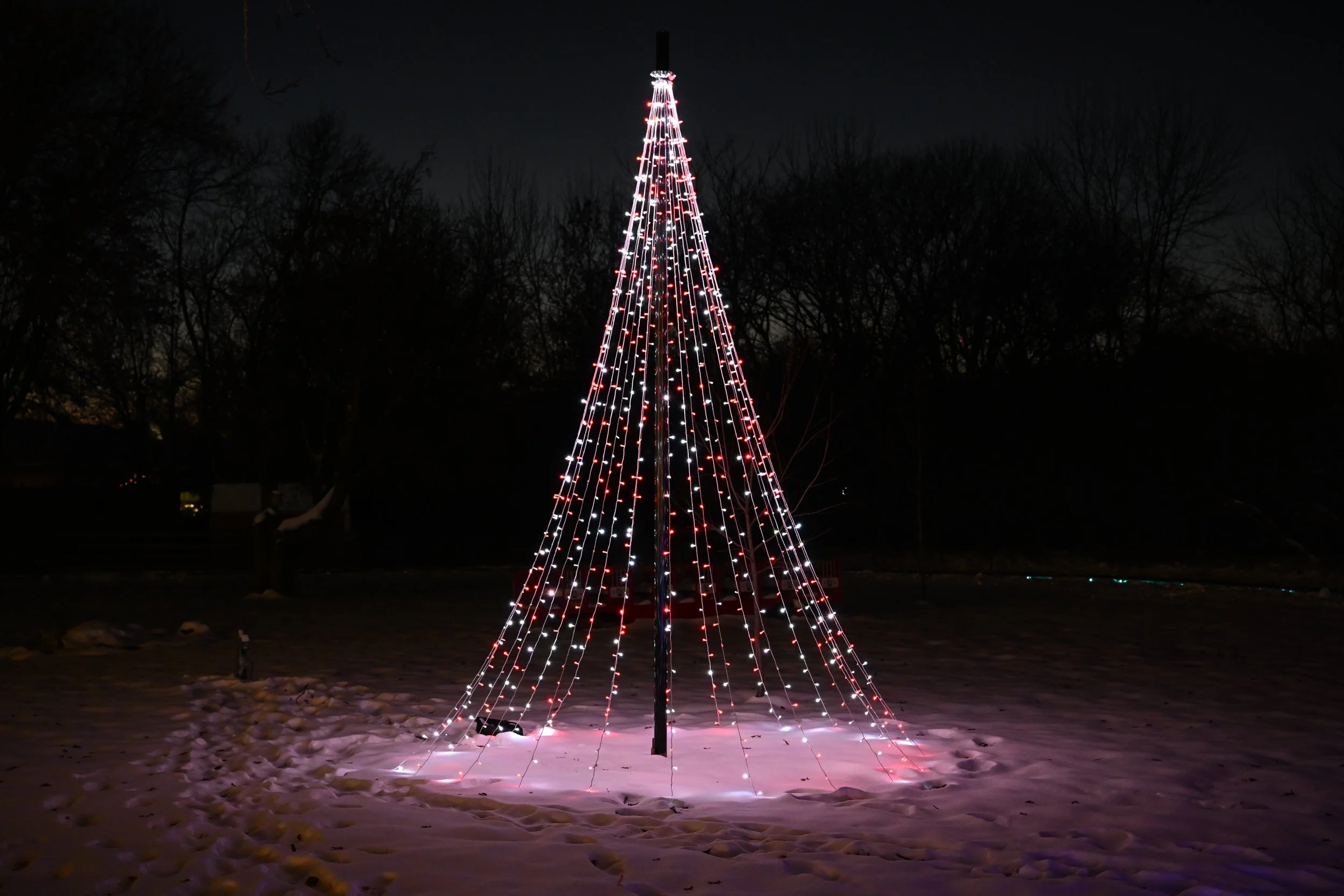 Christmas tree-shaped light display with white and red lights on a black pole in a snowy outdoor area at night.
