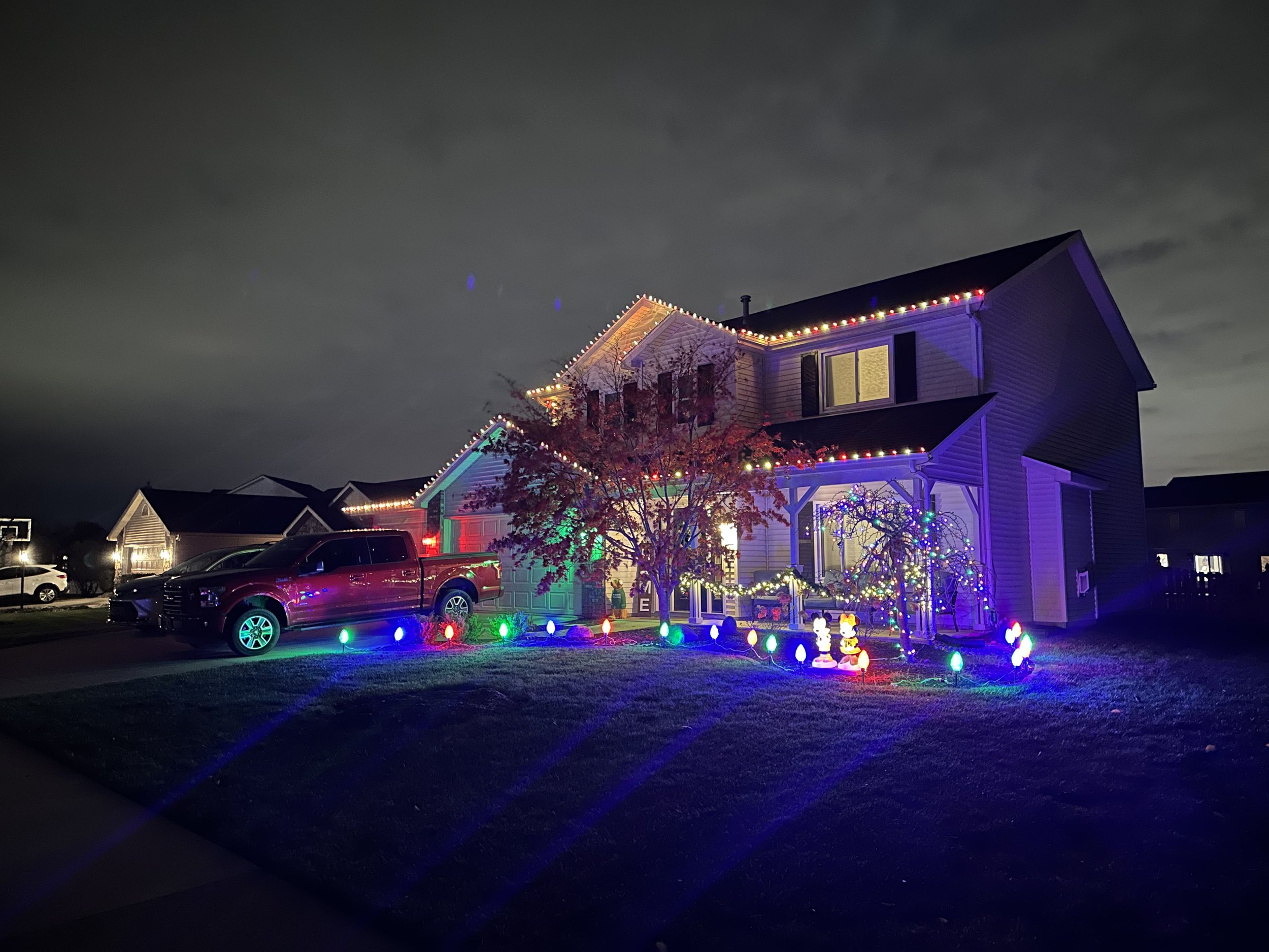 A two-story house decorated with colorful Christmas lights, including strings along the roofline and illuminated windows. There are lit decorations on the lawn, including a tree with lights and festive figures. Several cars are parked in the driveway, and the sky is dark with clouds.