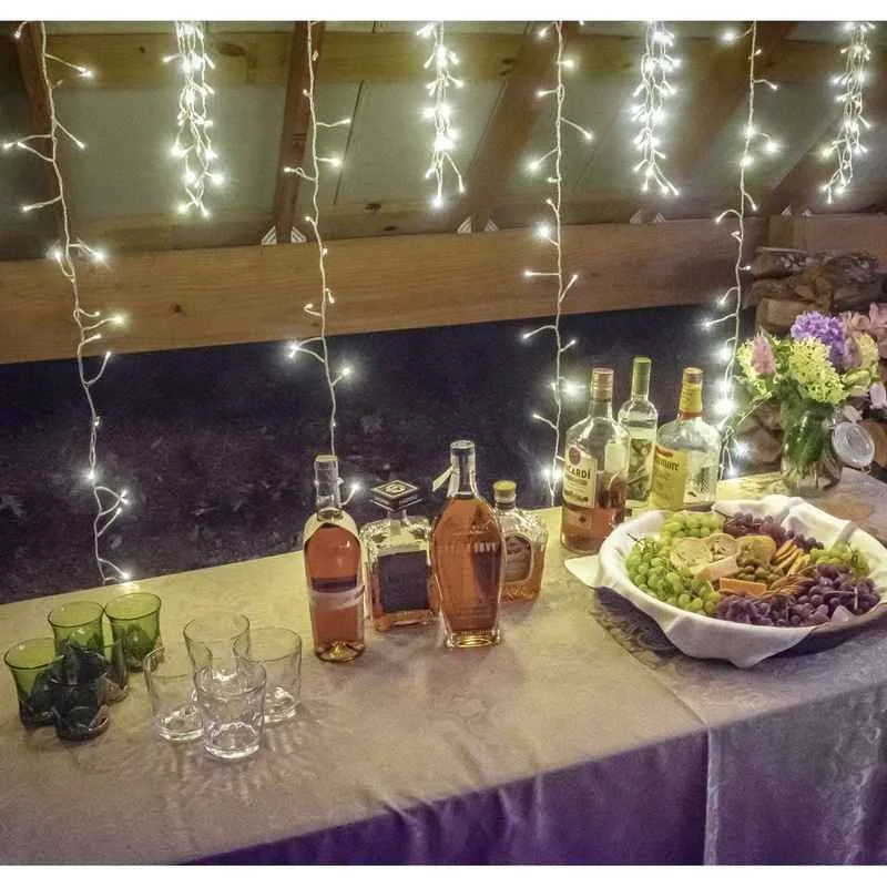 A table with bottles of alcohol, glasses, a large tray of grapes and cheese, and a vase with flowers, under string lights in a rustic setting.