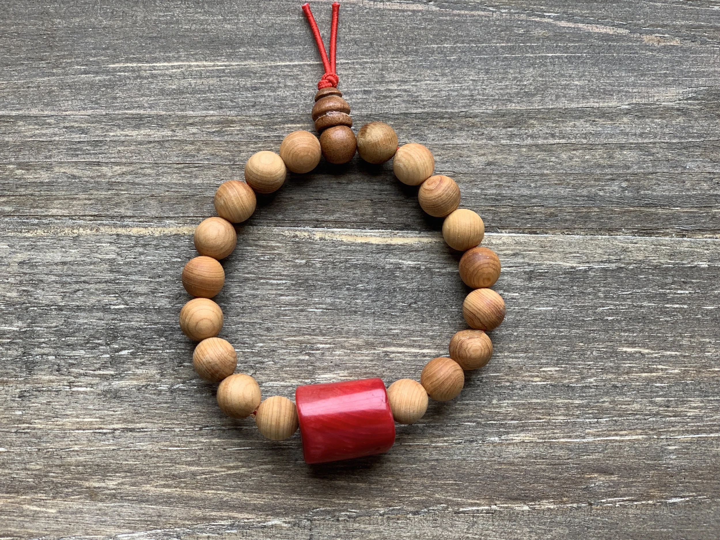 A beaded necklace with round wooden beads, a larger red cylindrical bead, and a red string on a wooden surface.