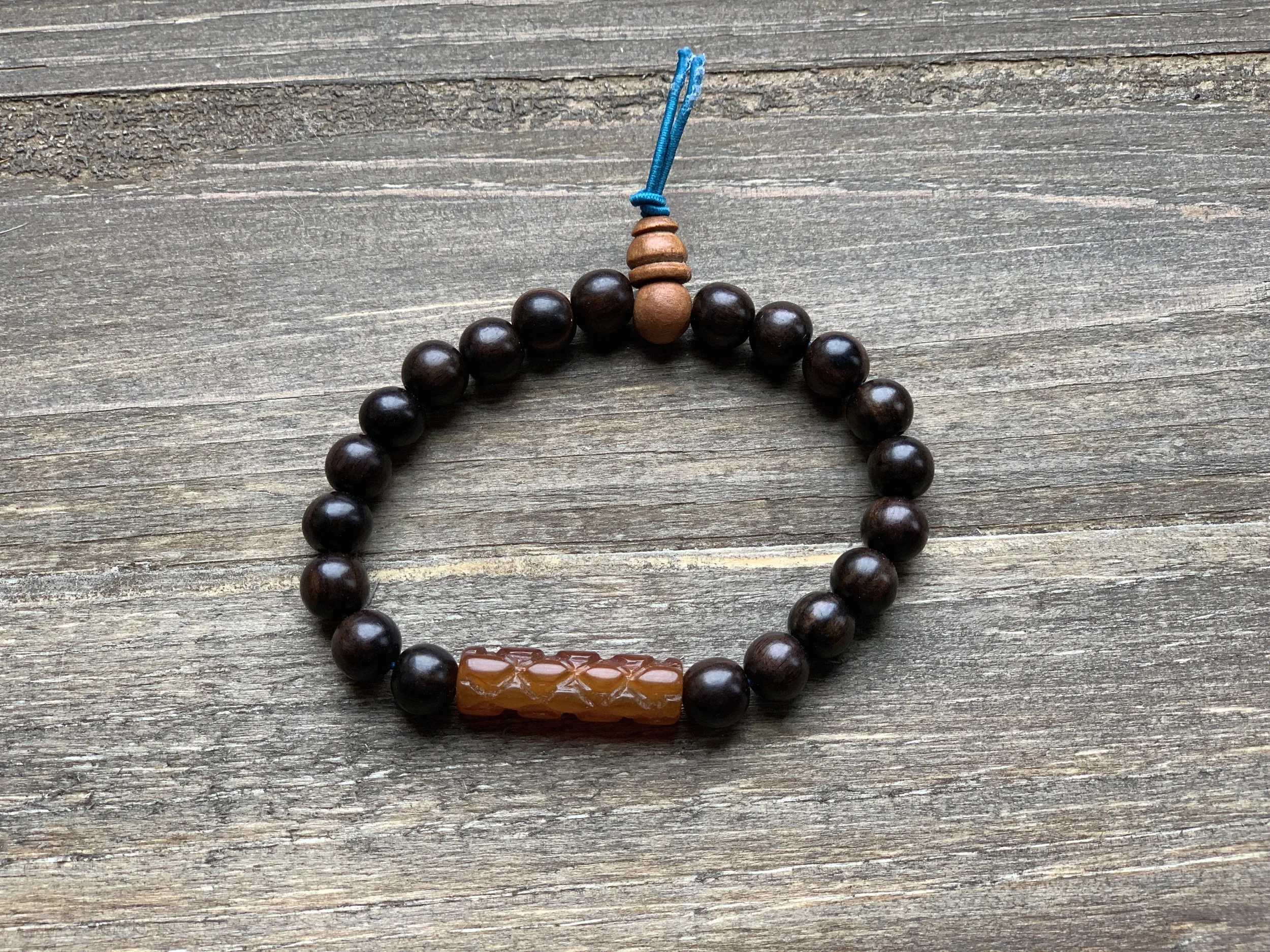 A beaded bracelet with dark brown beads and a carved orange bead, with a small blue cord at the end, placed on a weathered wooden surface.