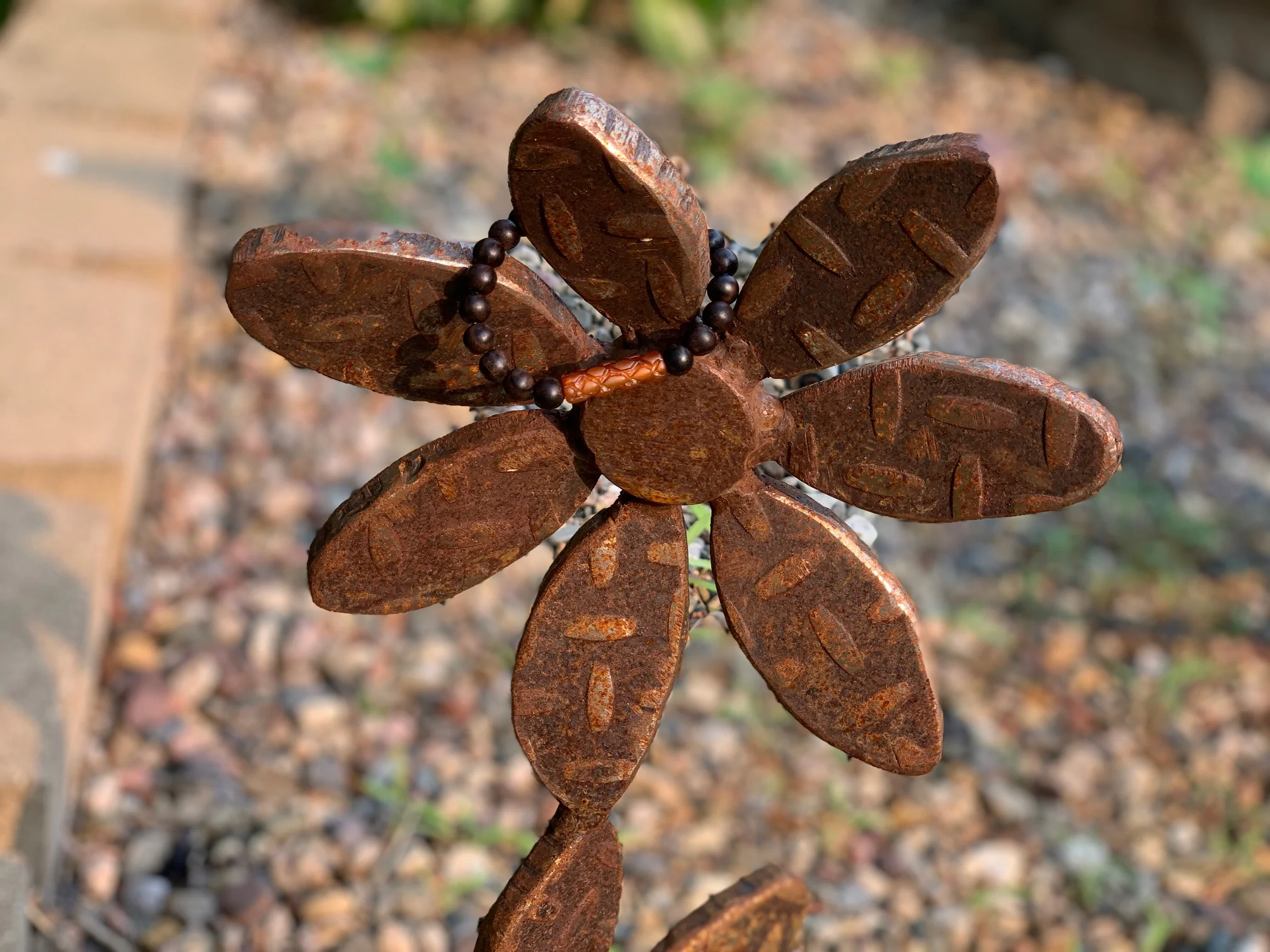 A close-up of a decorative metal flower sculpture with rusted petals, adorned with a black bead necklace, against a blurred outdoor background.