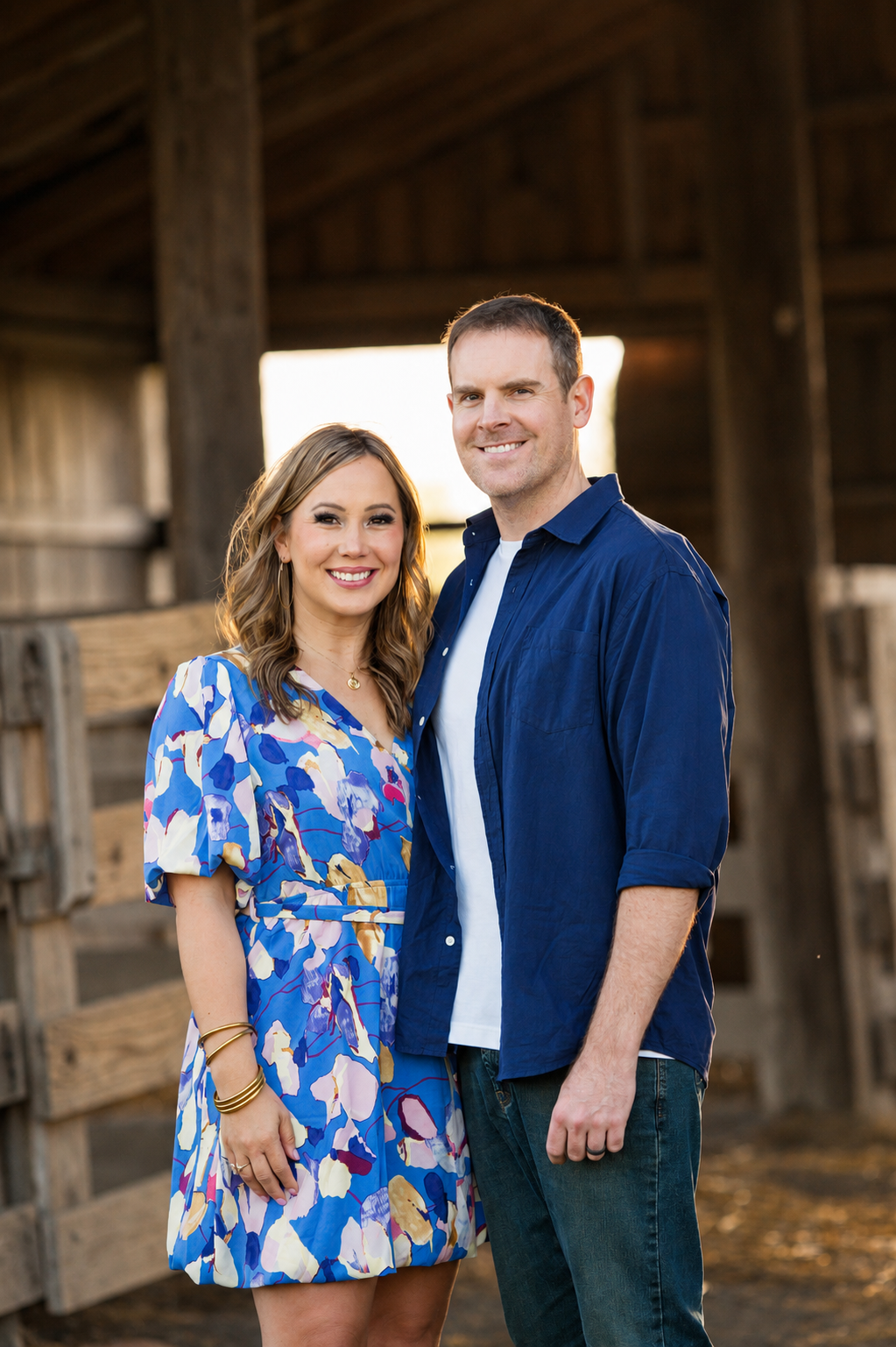 A smiling couple standing close together inside a rustic barn, with the woman wearing a colorful floral dress and the man in a blue button-up shirt and white T-shirt, during sunset.