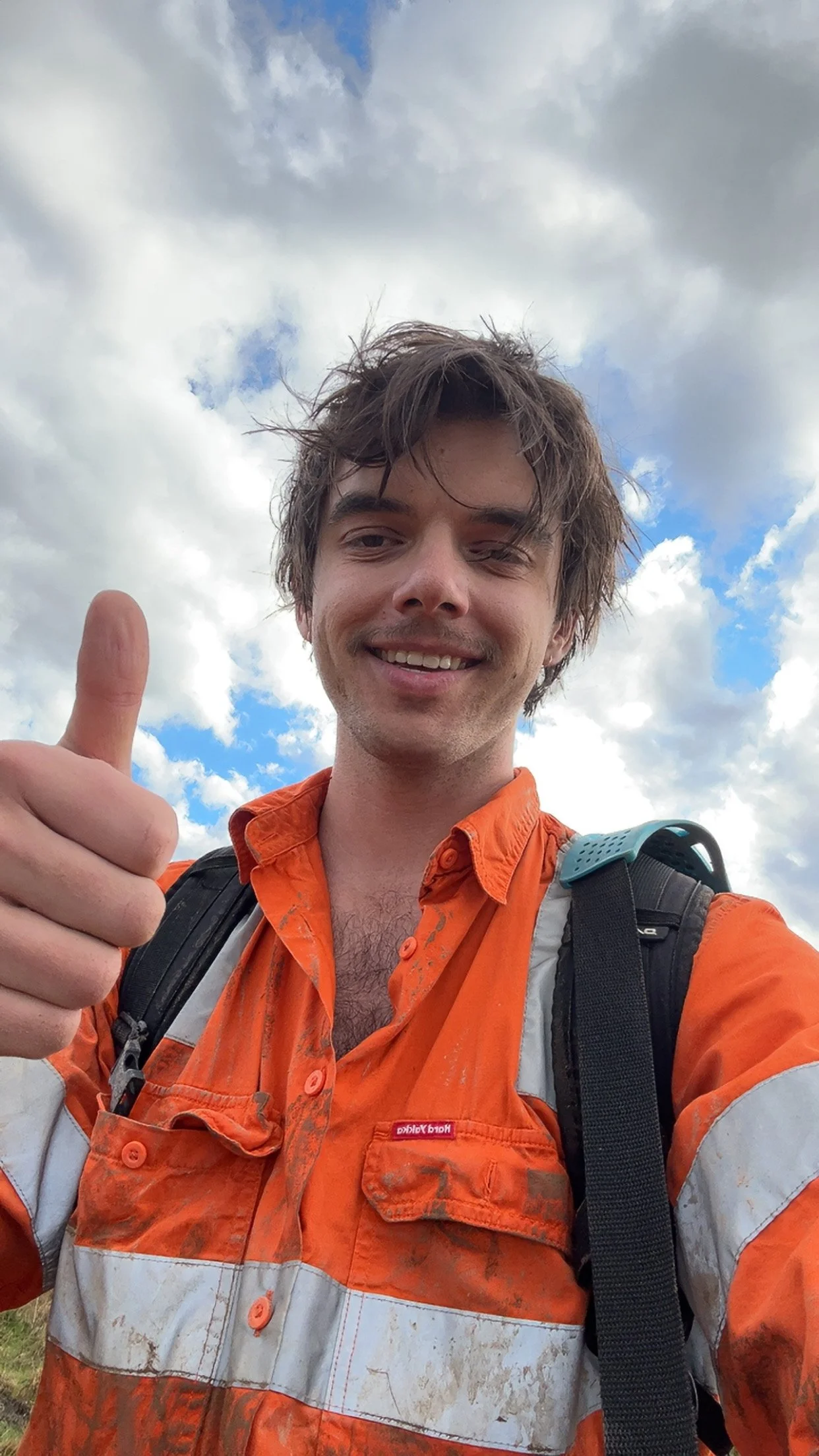 A selfie of Ryan at what looks like the end of a long day. His hi-vis shirt is dirty, and he looks tired, but he is smiling and giving us a thumbs-up. He is framed against a beautiful cloudy blue sky.