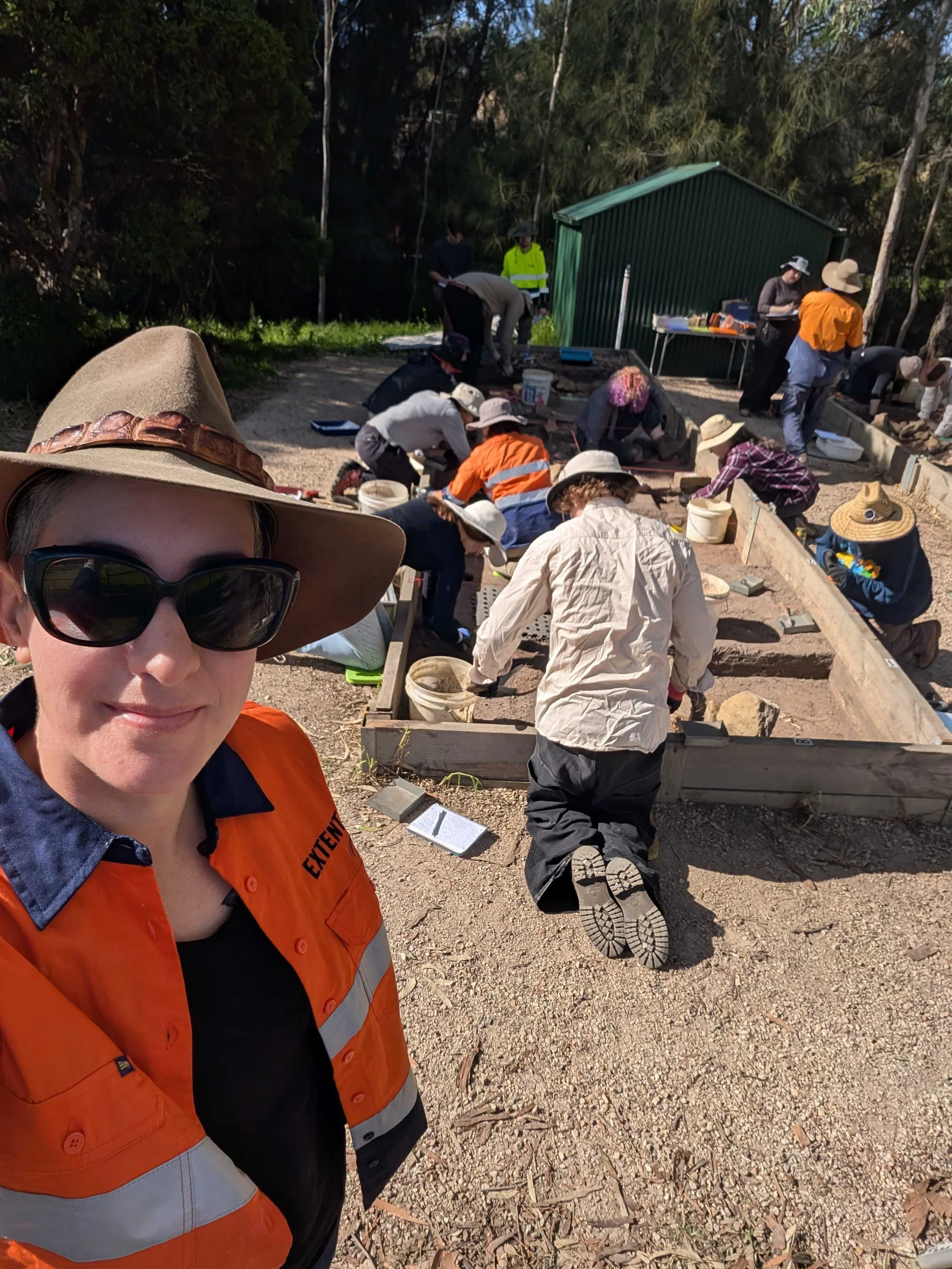 A selfie of Erica, wearing a broad-brimmed hat, sunglasses and a hi-vis shirt. In the background, a group of people are busily excavating and recording a mock archaeological site.
