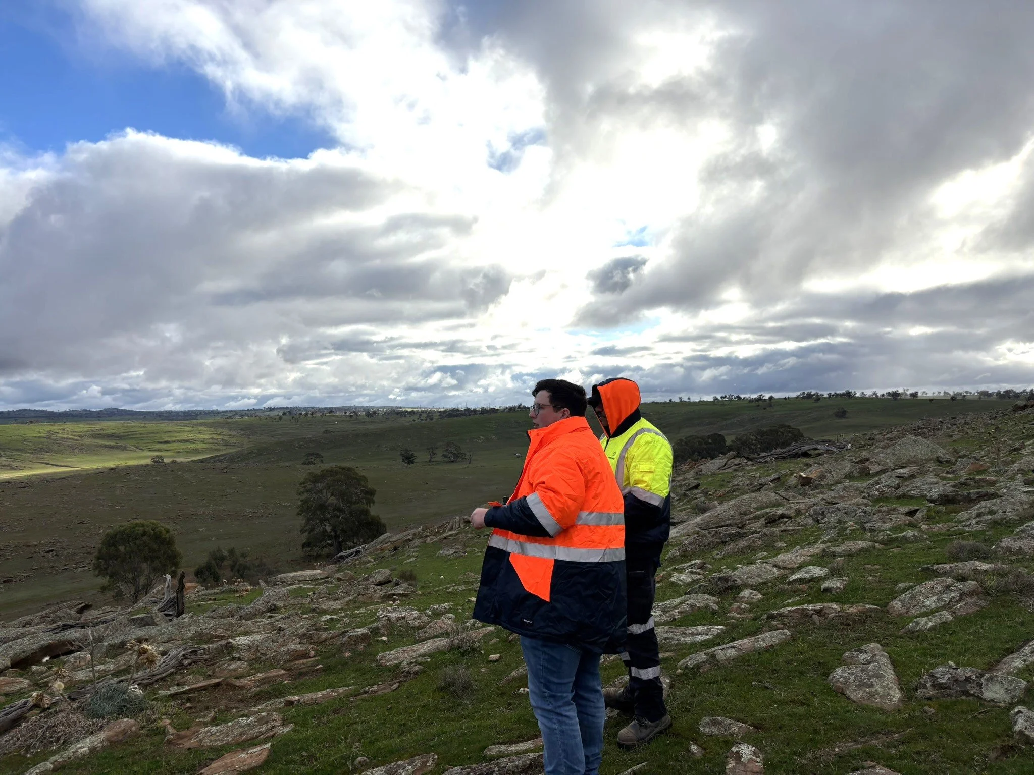 Cameron and a colleague are standing on the slope of a hill, looking over a landscape of rolling hills with outcropping rock. The sky is blue but cloudy, and it looks cold. They are wearing bulky hi-vis jackets.
