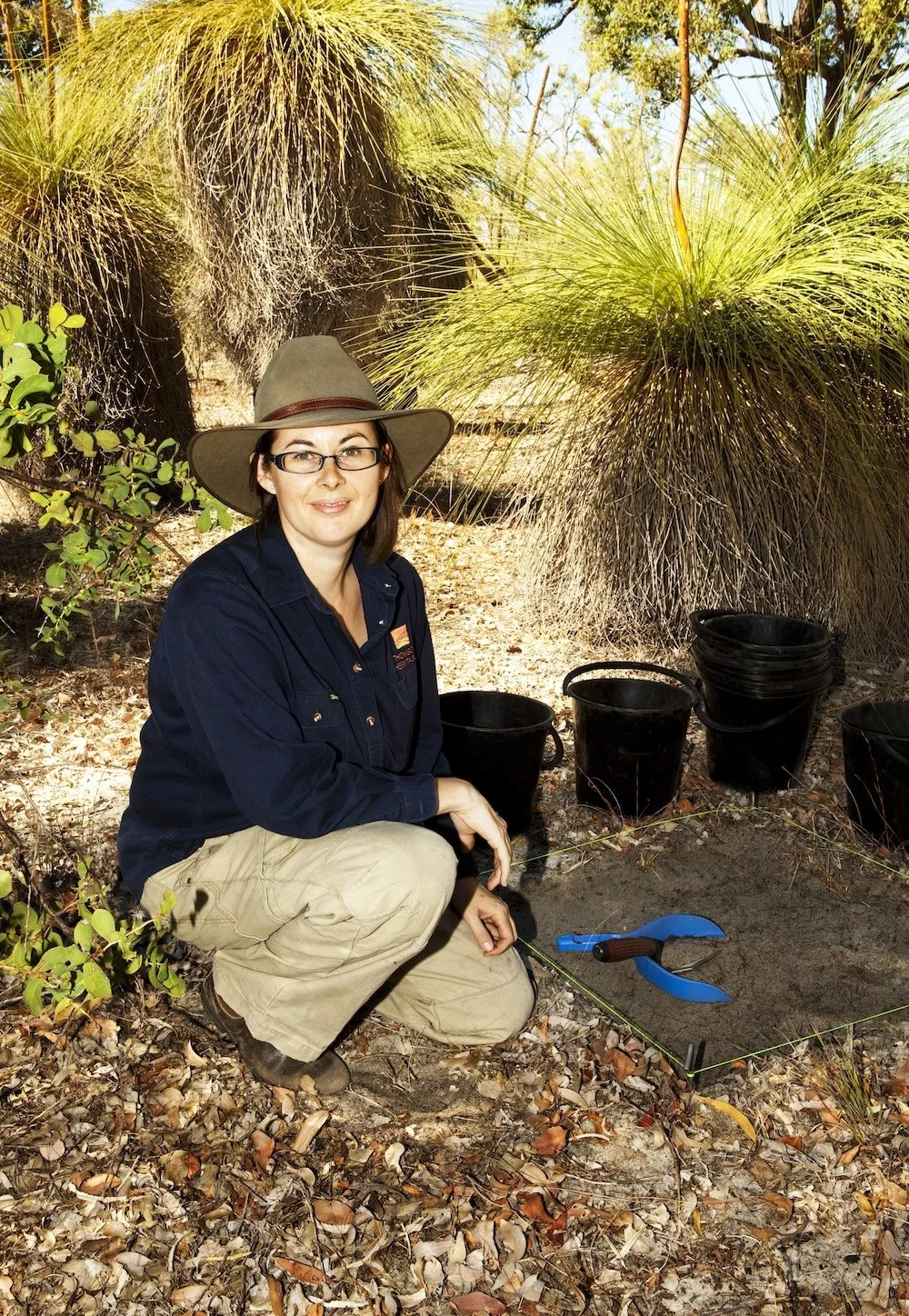 Jo is crouching on the ground, with a stand of grass trees in the background. She has just strung out a one-metre pit and has started excavation.