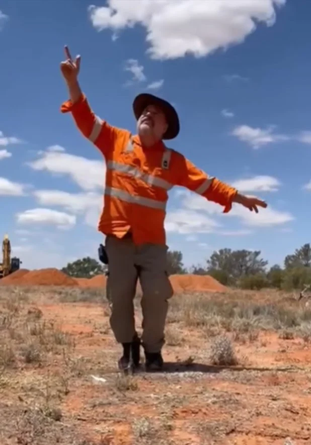 Michael, wearing a hi-vis shirt and work-wear, is silhouetted against a blue sky with patchy clouds, dancing on the red dirt.
