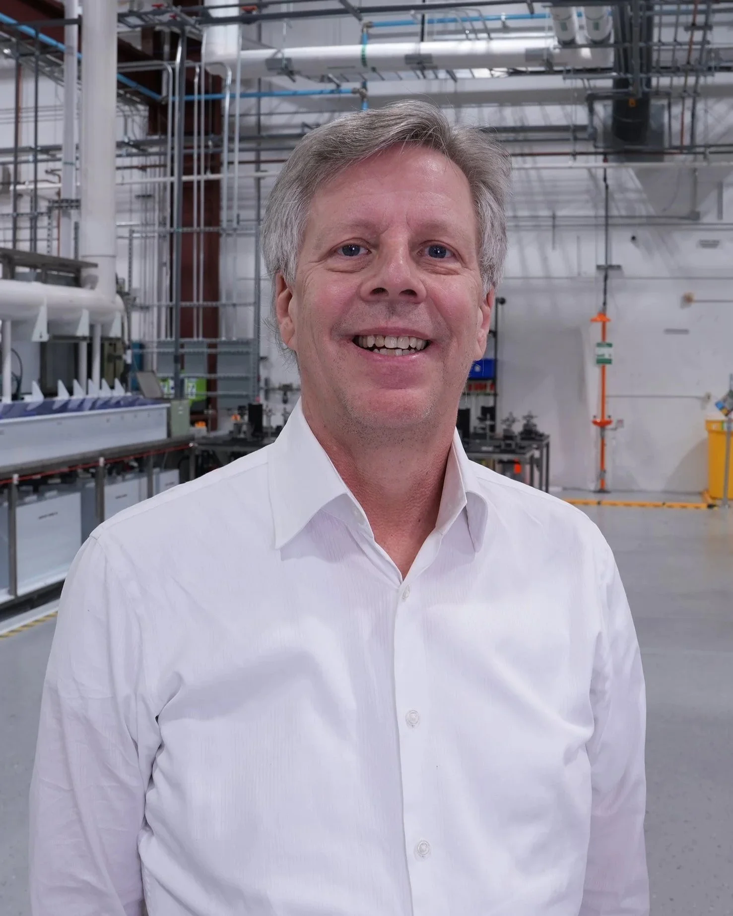 A man in a white shirt smiling in an industrial or laboratory setting with metal pipes and equipment in the background.