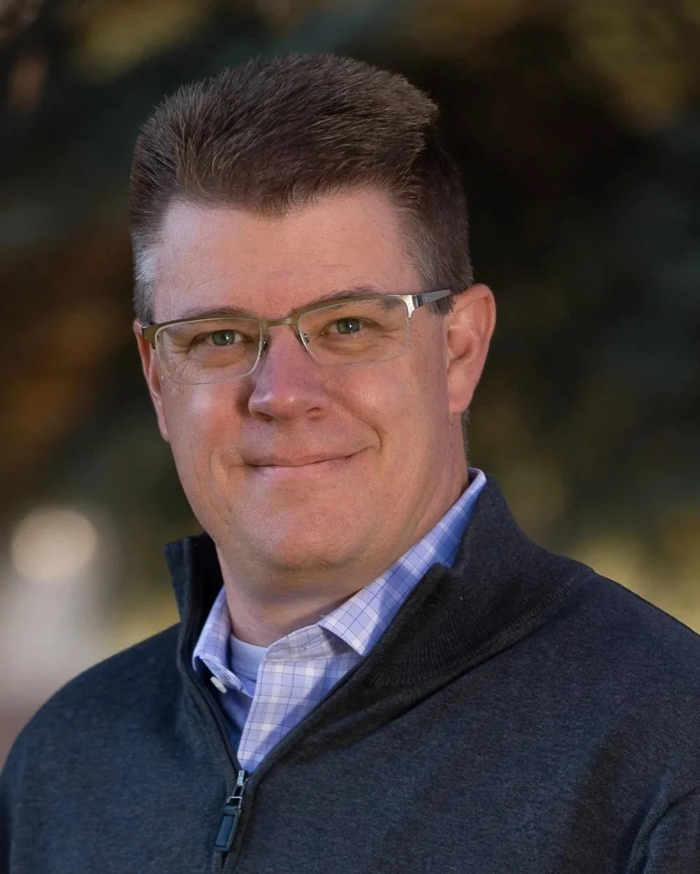 Headshot of a man wearing glasses, a collared shirt, and a zip-up sweater, smiling outdoors.
