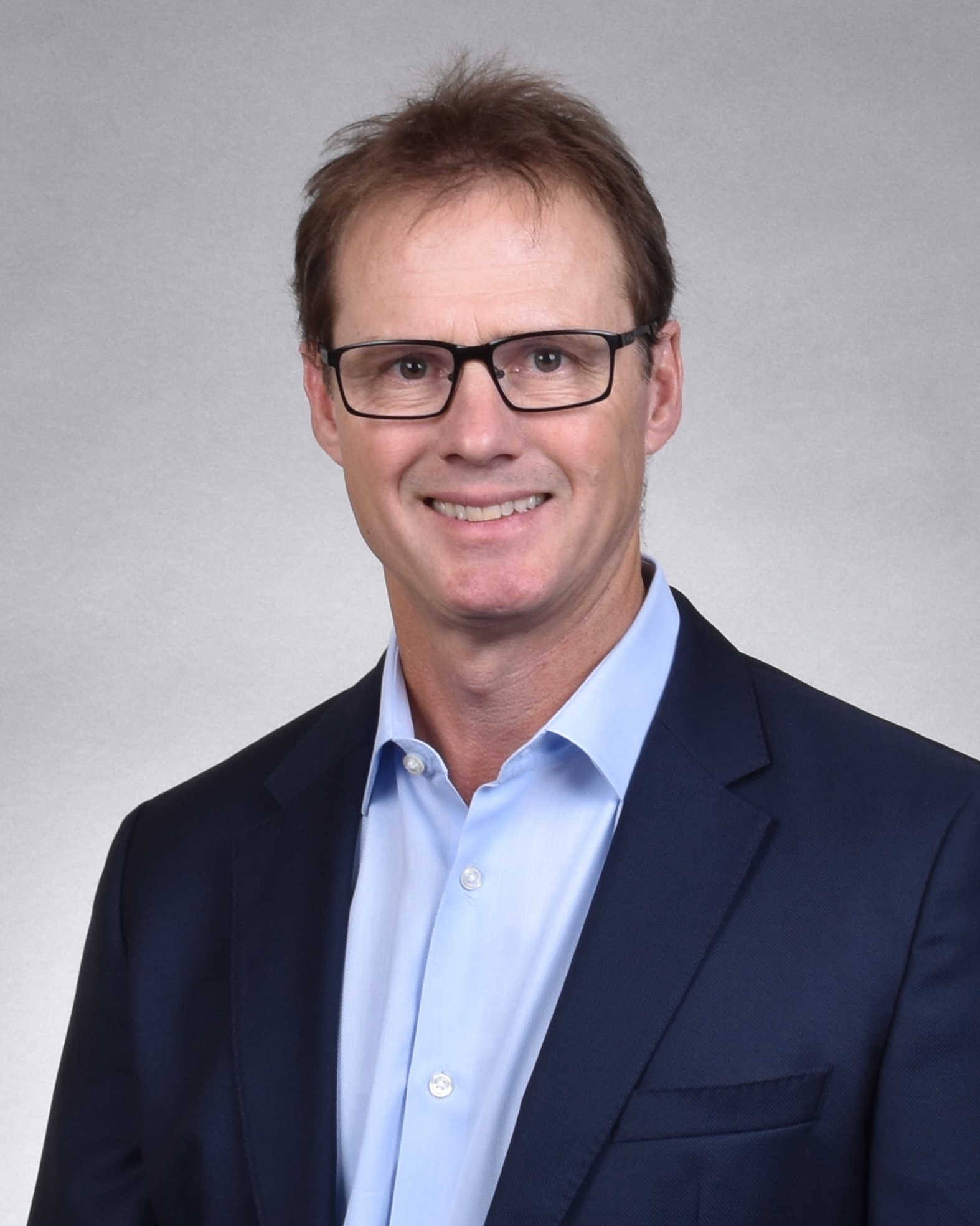 Professional headshot of a man wearing glasses, a light blue shirt, and a dark blazer, smiling against a neutral background.