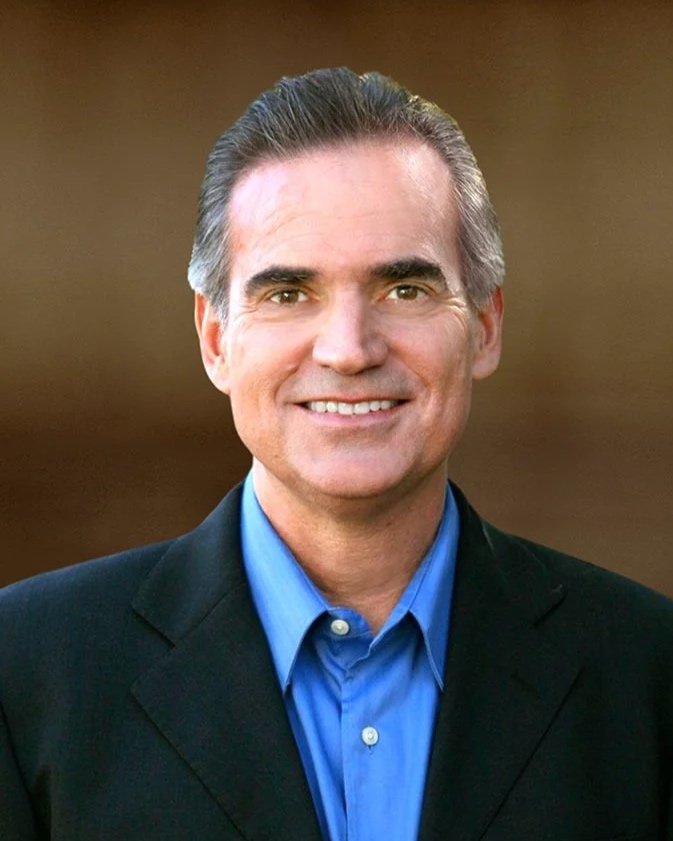 A middle-aged man with neatly combed gray hair, wearing a dark suit jacket and a blue collared shirt, smiling at the camera with a blurred brown and beige background.