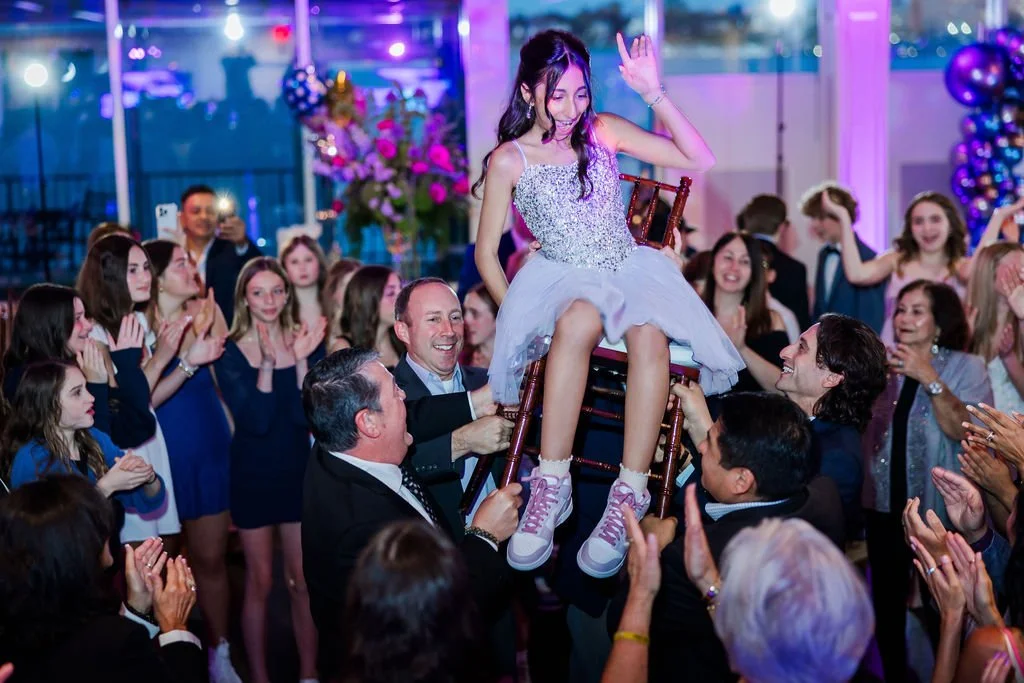 Girl in a sparkly dress being lifted on a chair while people cheer and celebrate at a party.