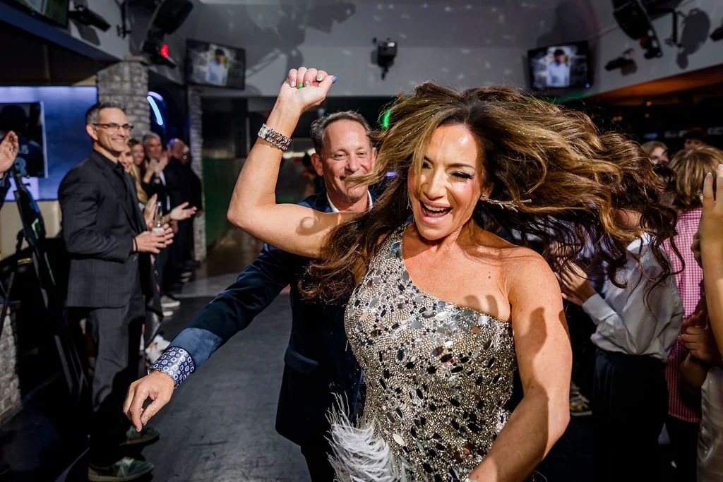 A woman with long brown hair dancing with joy at a party, wearing a sparkly silver dress, surrounded by people clapping and smiling, in a lively indoor setting.