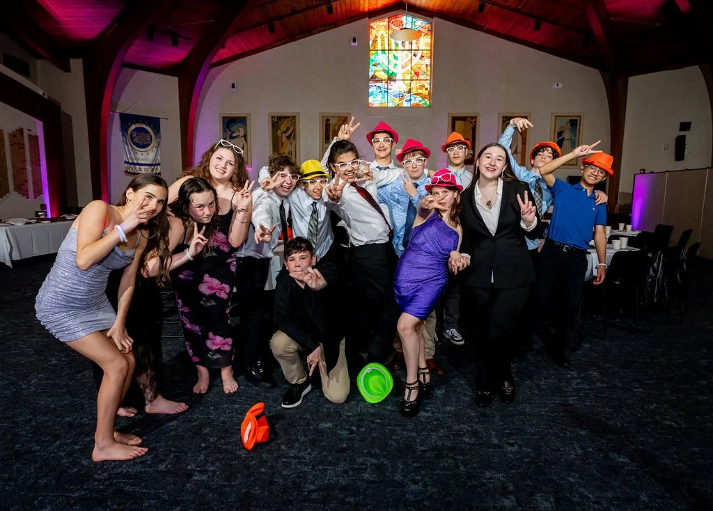 Group of young people dressed in casual and semi-formal attire posing and making fun gestures at a party or gathering inside a venue with high ceilings, stained glass window, and colorful lighting.