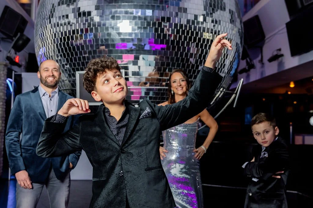 A group of four well-dressed people at a party or club, with a large disco ball overhead. A boy in the foreground is dancing, while a woman in a silver dress and two men are standing and smiling in the background.