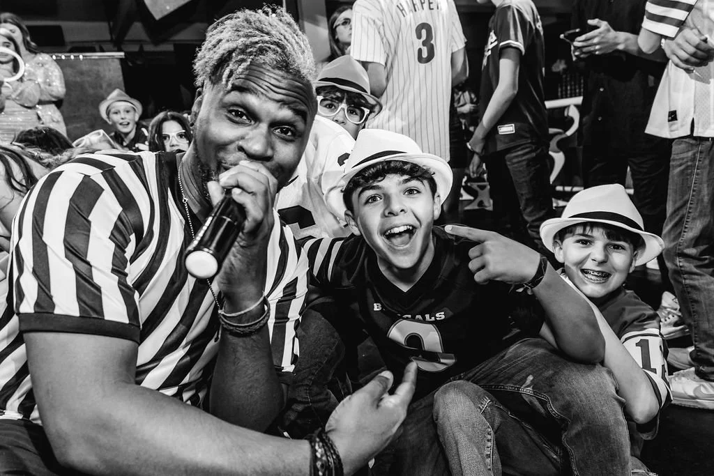 A man with a microphone and dreadlocks, wearing a striped shirt, poses with three young boys wearing hats and football jerseys at a lively event with other people in the background.