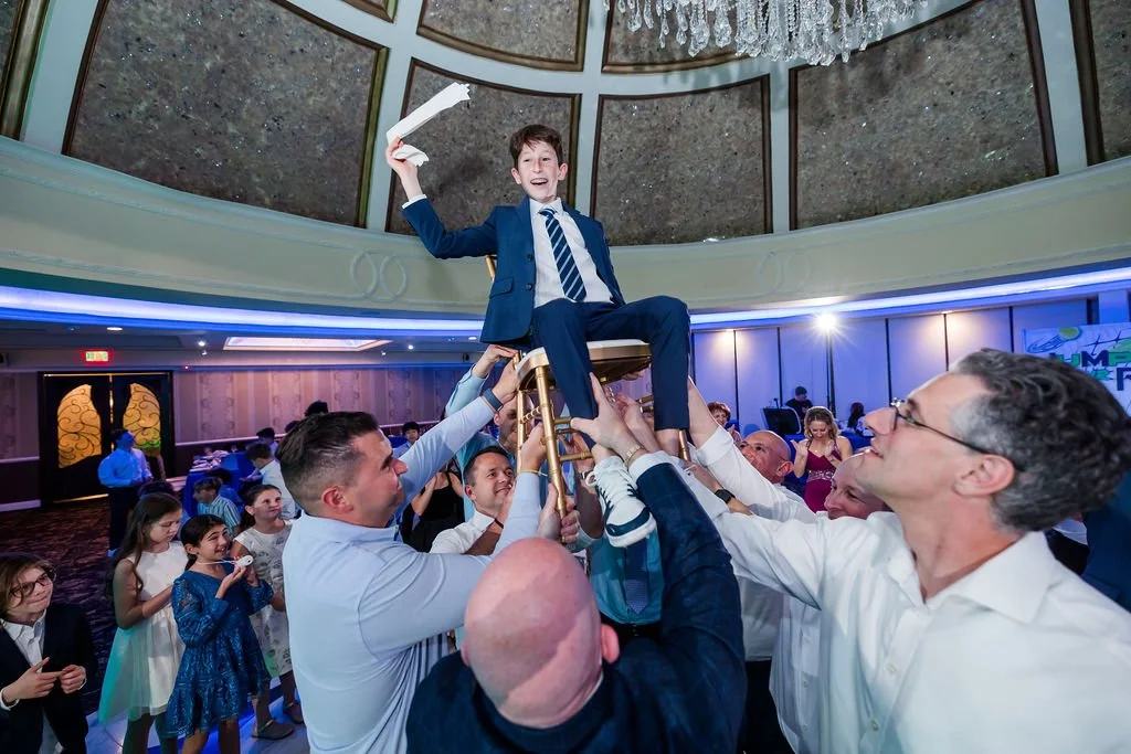 Group of men lifting a young boy in a chair during a celebration or party, with children and adults watching.