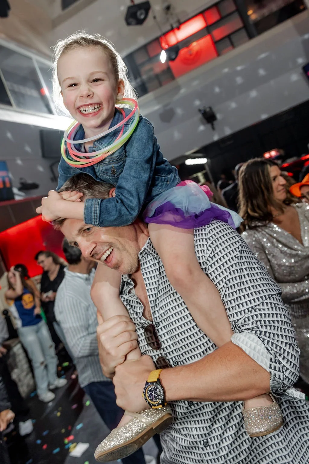 A smiling young girl sitting on a man's shoulders at a lively party or celebration, with colorful necklaces and a purple tutu. The man is holding her legs, and both appear happy. Other people are dancing and socializing in the background.