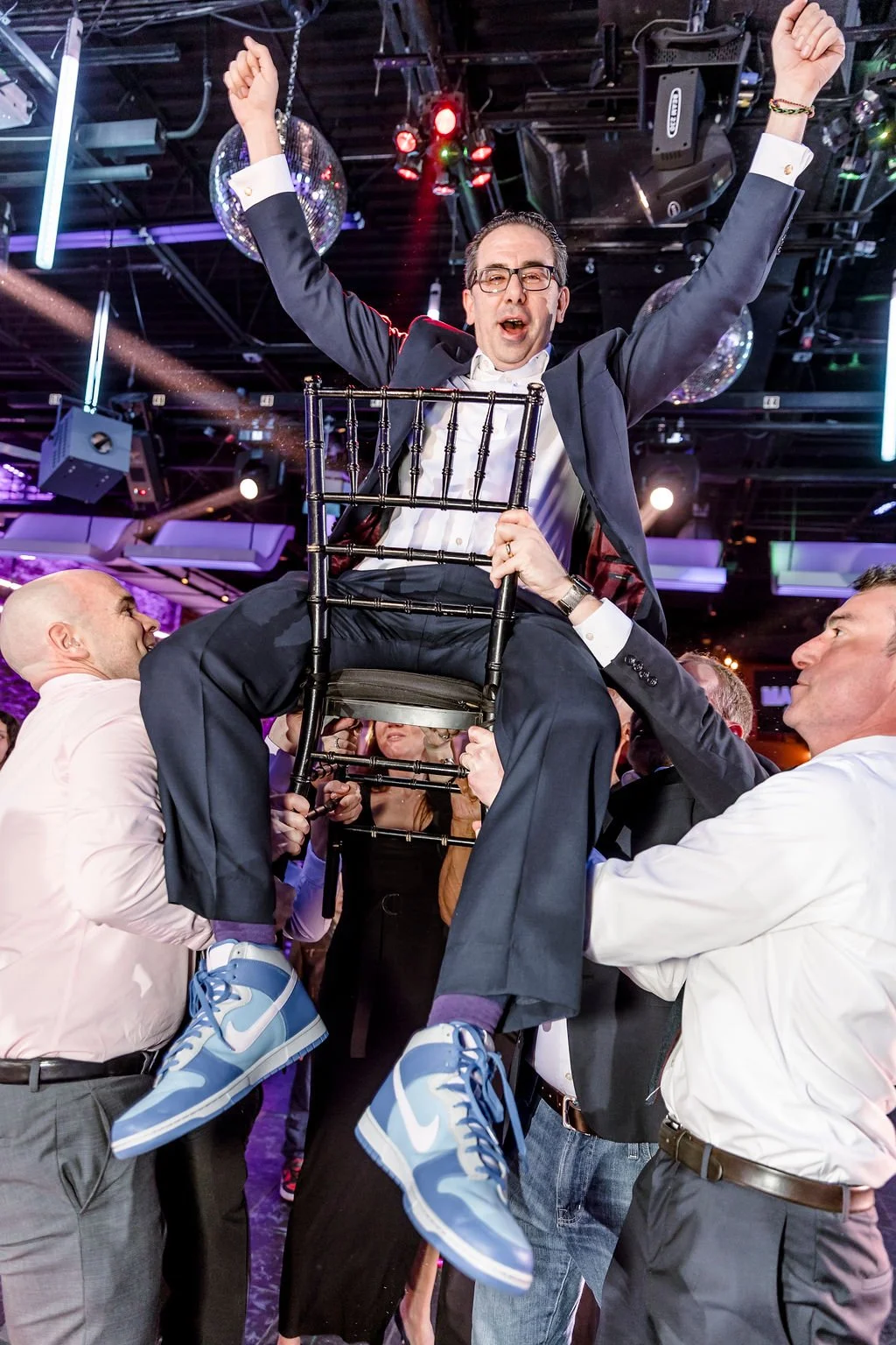 A man dressed in formal attire sits on a chair held up by several people during a celebration or party in a lively, colorful venue with disco lights.