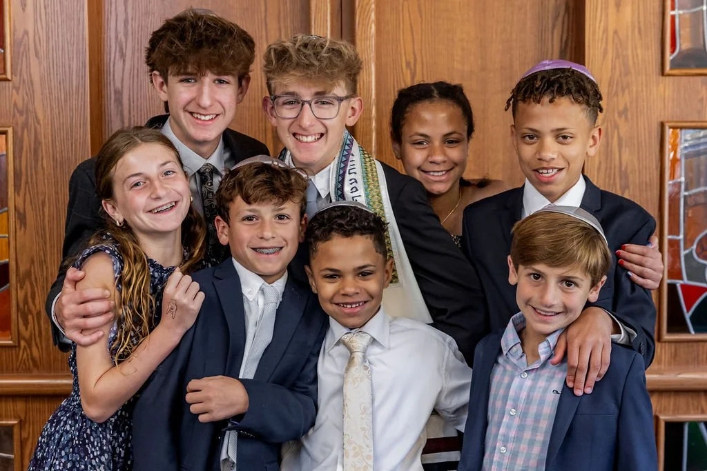 Group of nine children smiling and posing for a photo inside a wooden-paneled room, some wearing suits and a girl in a floral dress, with one boy wearing a kippah.