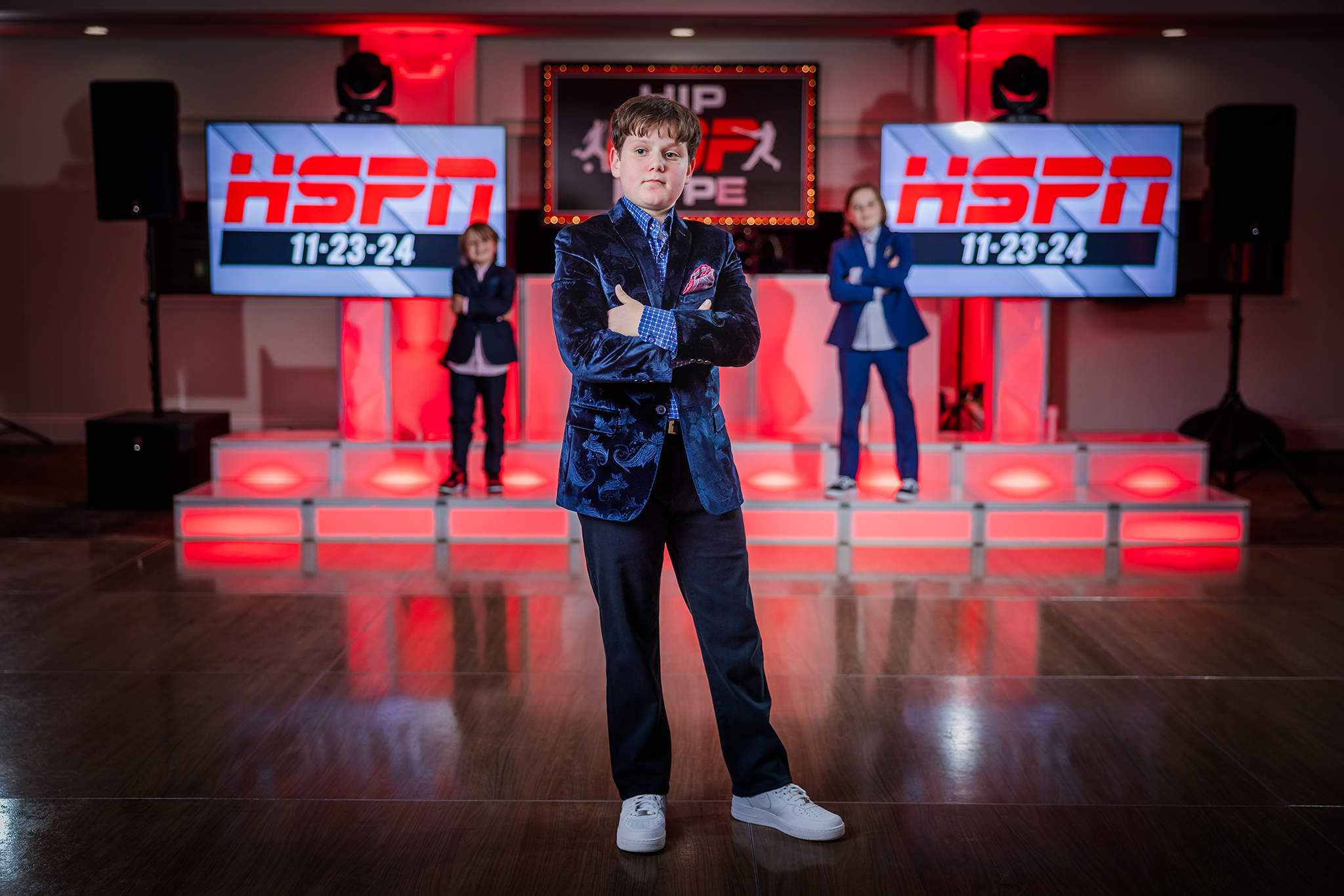 A young boy in a velvet blazer posing with crossed arms on a dance floor, with a stage in the background featuring two women in suits and large screens displaying the ESPN logo and the dates 11-23-24.