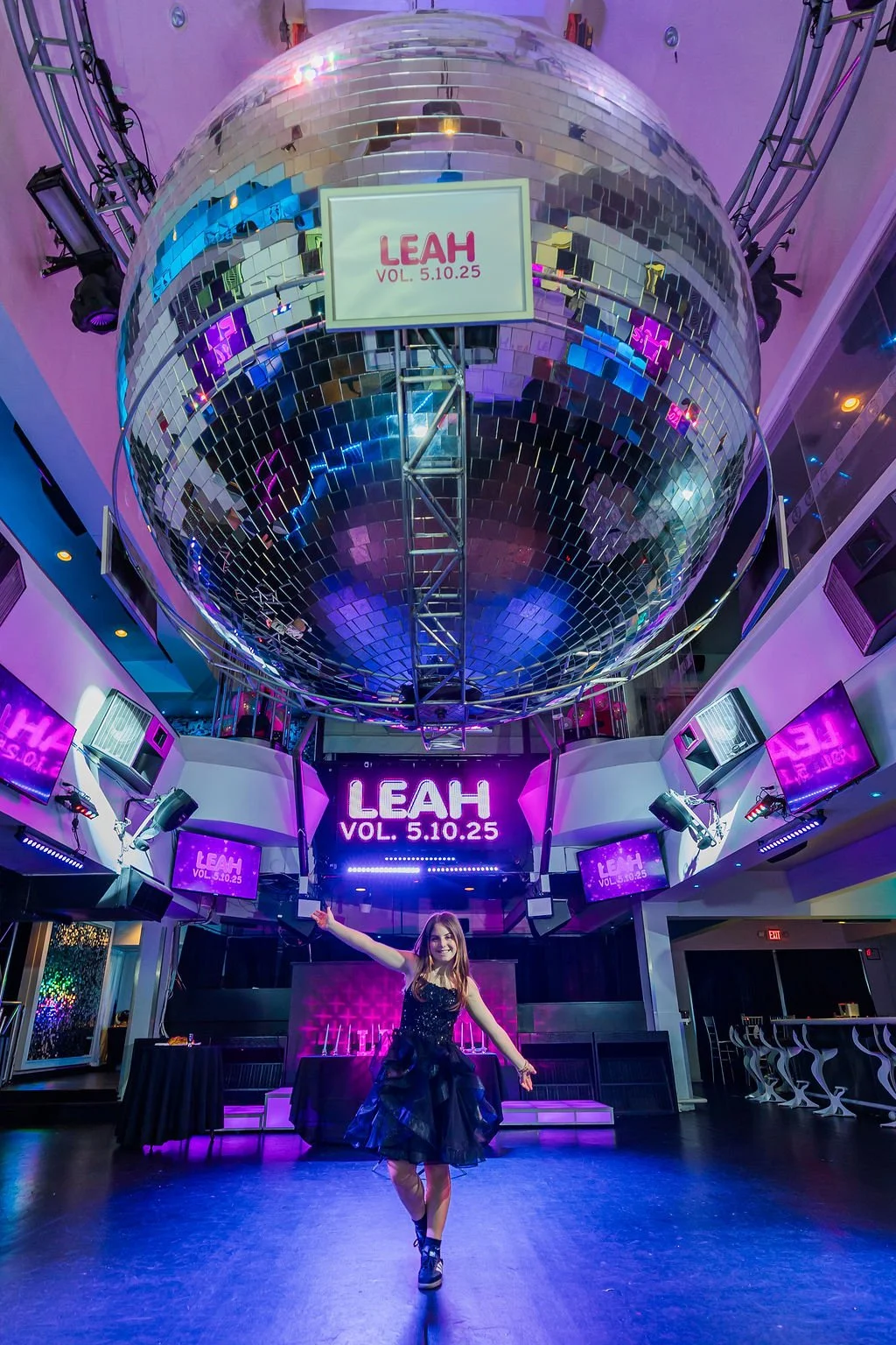 A woman in a black dress is dancing on a stage with purple neon lights saying LEAH VOL. 5.10.25 behind her. The stage is inside a nightclub with a mirrored disco ball hanging from the ceiling.