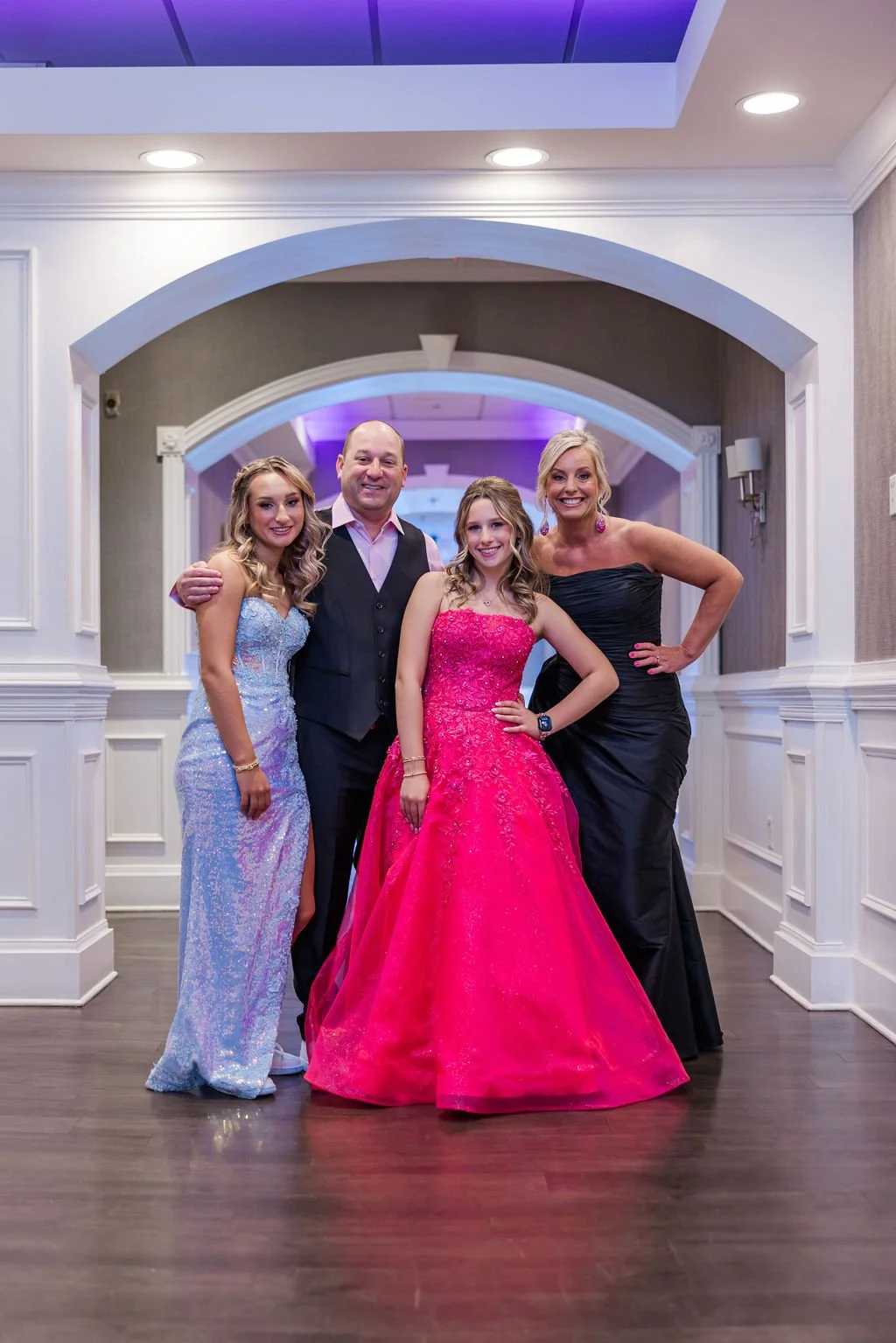 Group of four people dressed in formal gowns and suit, posing together at an indoor event. The young woman in pink is at the center, flanked by a man in a vest and shirt, and two women in black and light blue dresses.