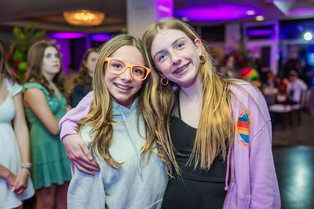 Two smiling girls with braces pose together at a party, with other girls in the background.