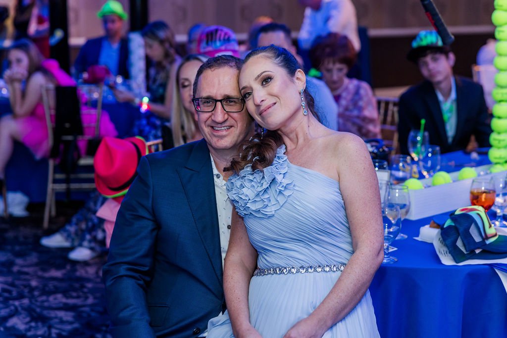 A couple in formal attire, smiling and sitting close together at a party or celebration, with children and other guests in the background.
