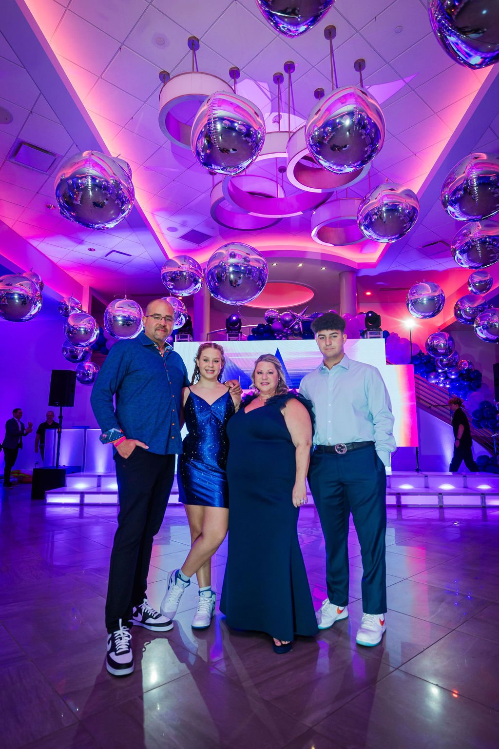 Group of four people posing at an event with purple and pink lighting, hanging silver balloons, and a stage with digital screens in the background.