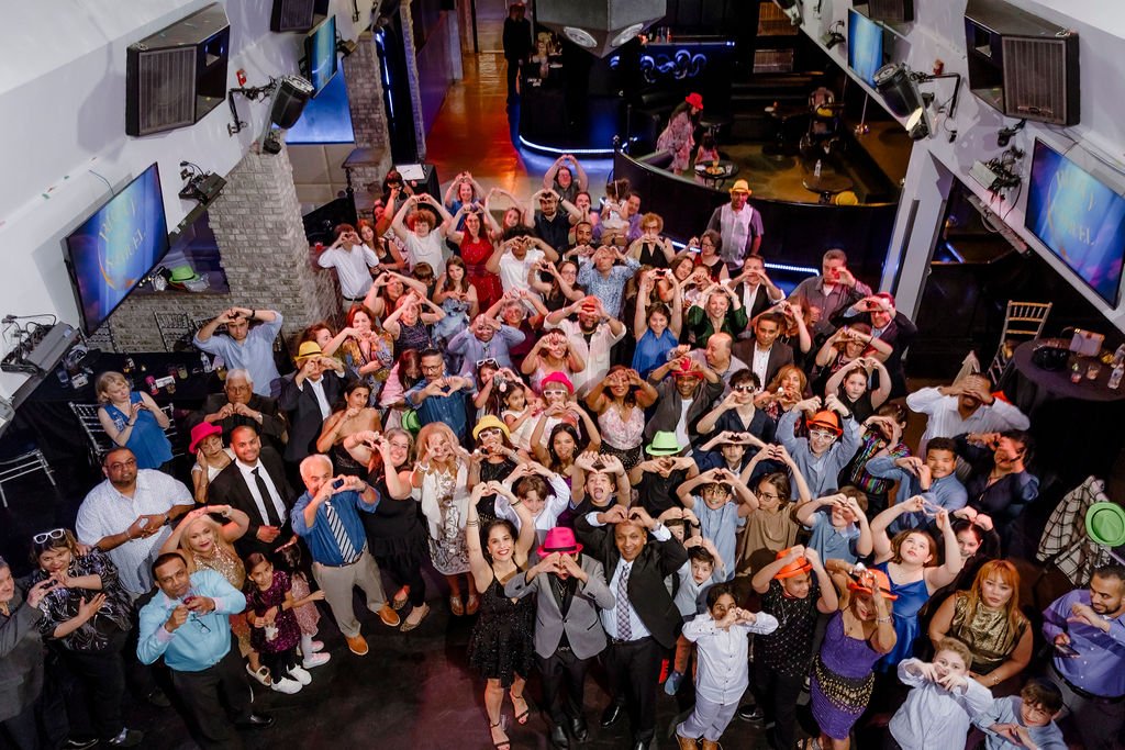 A large group of people in a dance club or event space, making heart shapes with their arms over their heads, some are wearing colorful hats, and there are multiple screens on the walls.