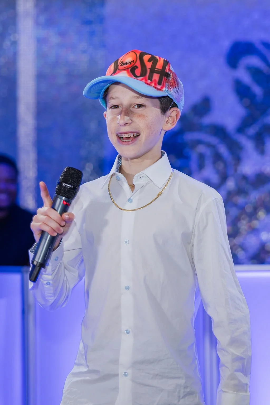 A young boy with short dark hair, wearing a white dress shirt, gold chain, and a colorful cap, is speaking into a microphone at an event with a blue background.