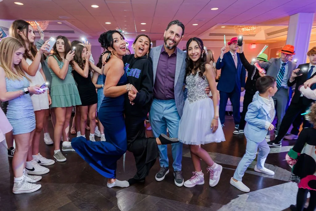 People dancing and celebrating at a party or event, with a group of four individuals in the center posing for a photo, surrounded by children and other attendees.