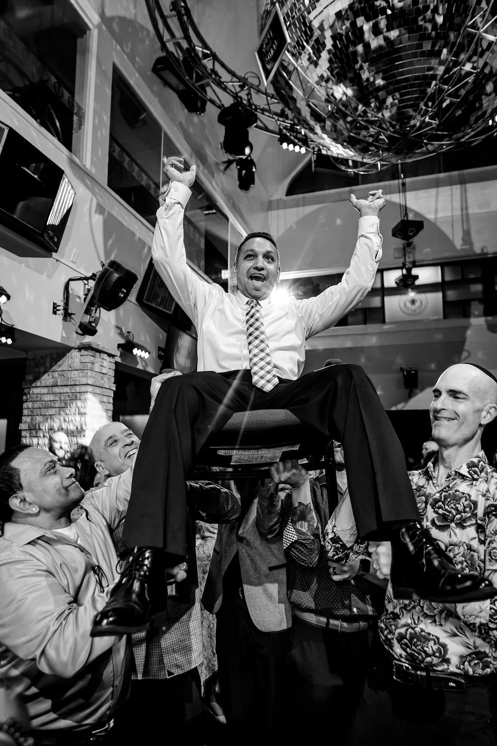 A man in a white shirt and striped tie is lifted in a chair by multiple people at a celebration event. He has his arms raised in victory and is smiling, surrounded by smiling supporters.