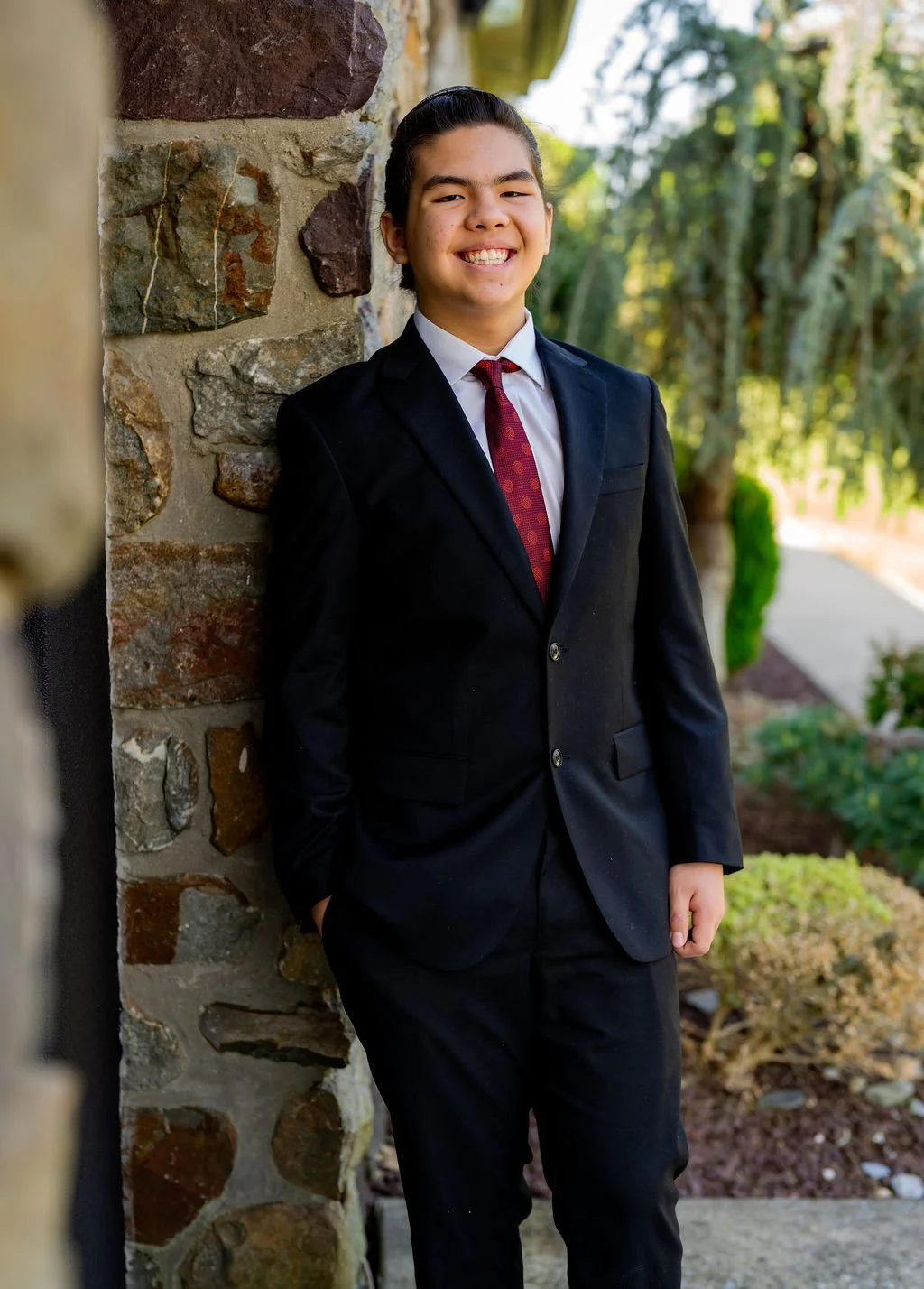 A young boy dressed in a black suit with a white shirt and red tie, smiling and leaning against a stone wall outdoors.