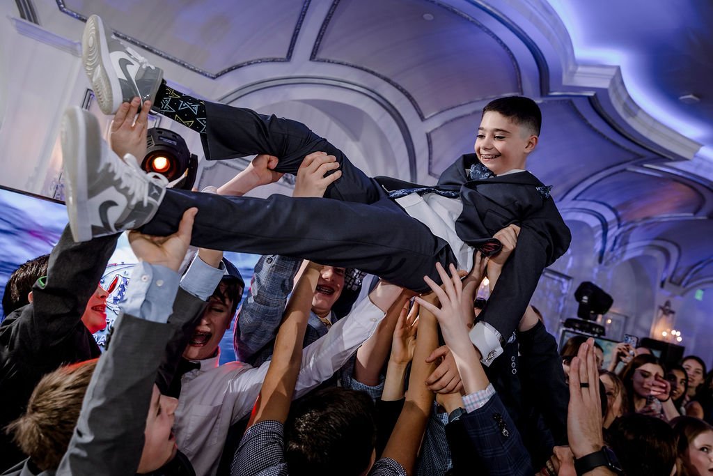 A young boy in a black formal suit is being lifted and celebrated by a crowd at a party, with joyful expressions and a festive indoor setting.