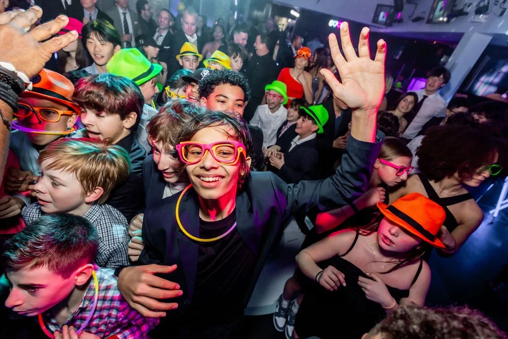 Group of children at a party wearing colorful hats, glasses, and glow sticks, dancing and having fun.
