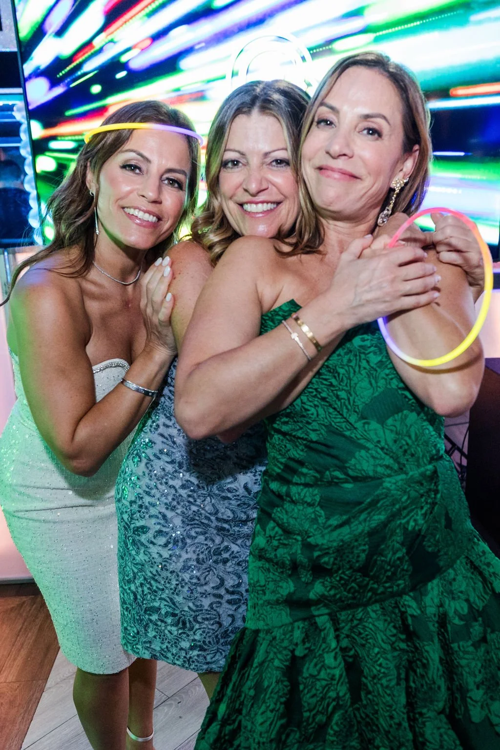 Three smiling women celebrating at a party, holding glow sticks, with colorful lighting in the background.