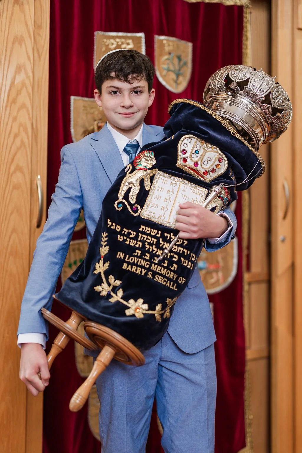 A young boy in a light blue suit holding a ceremonial Torah decorated with a crown and embroidered cloth, standing in front of a red curtain.