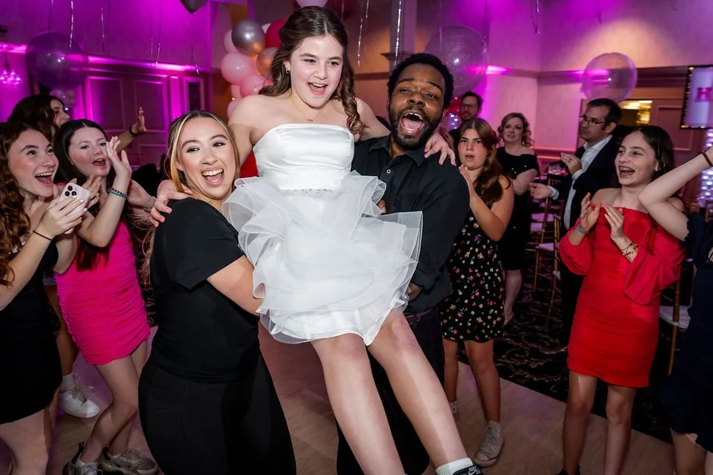 A young girl in a white dress being lifted by a man at a party with many young women surrounding them, some clapping and smiling in a pink and purple decorated venue.