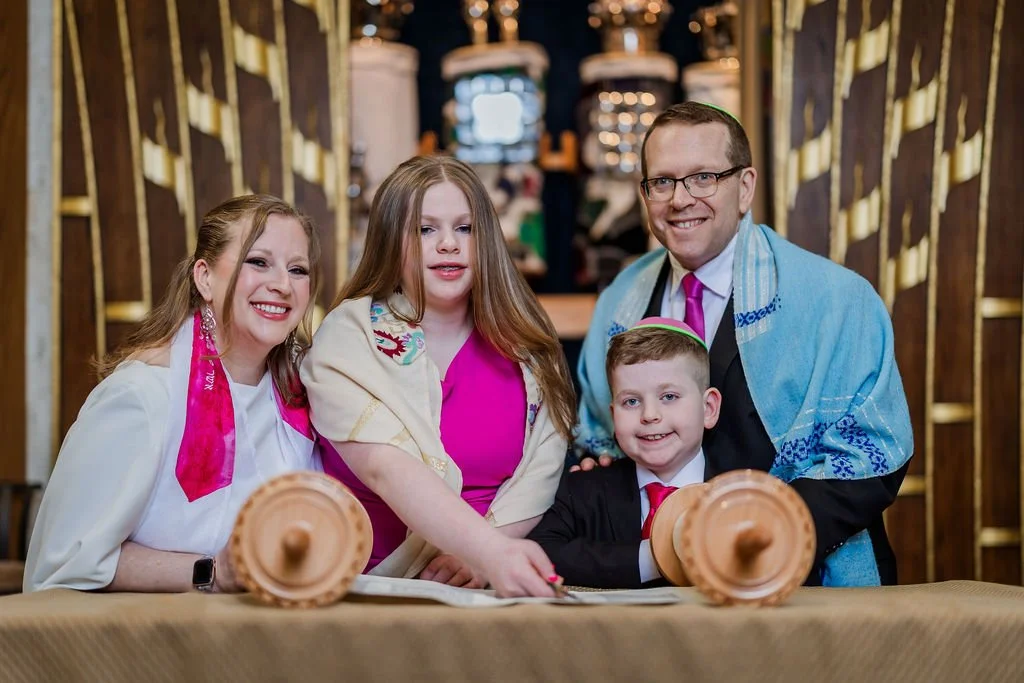 Family gathered around a table during a Jewish celebration, with a Torah scroll in the foreground, inside a synagogue.