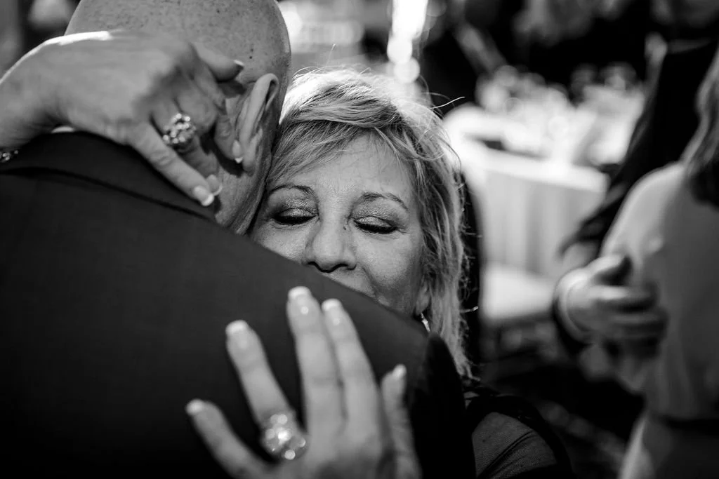 A woman with closed eyes hugging a man. The woman's hand rests on the man's shoulder, showing rings on her fingers. The scene is warm and emotional.