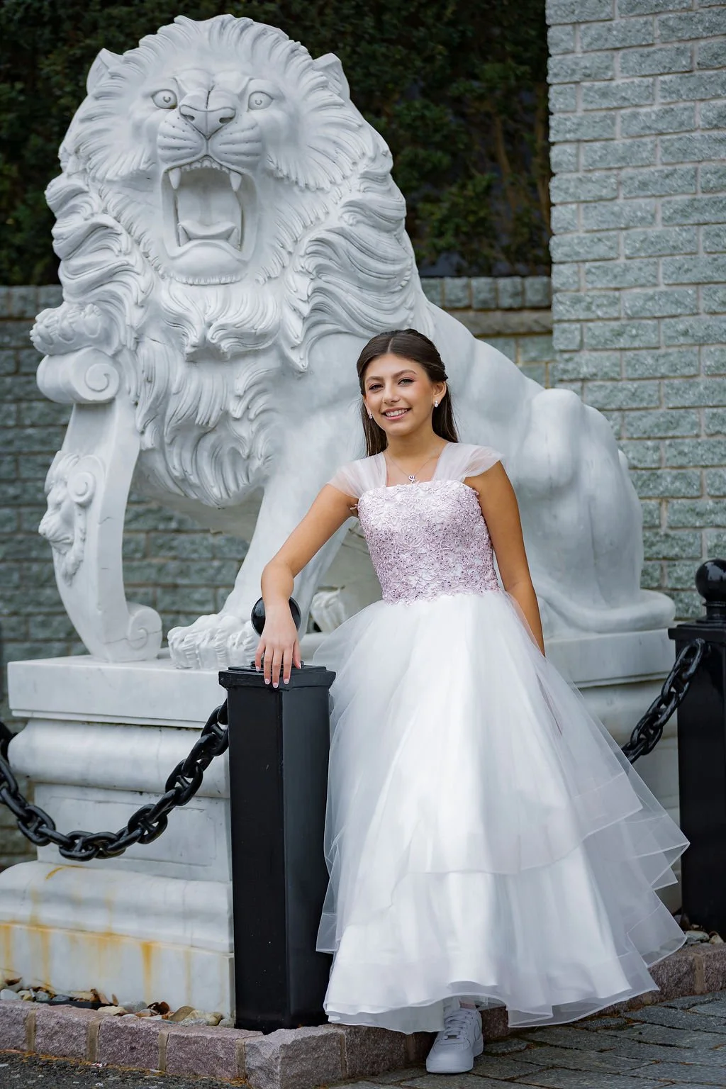 Young woman in a white wedding dress standing in front of a large white lion sculpture with a brick wall in the background.