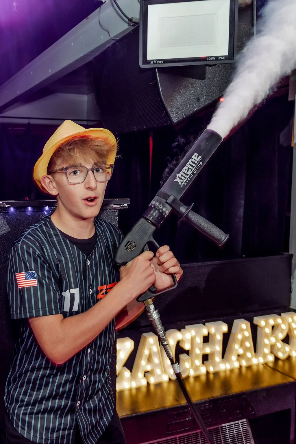 A young boy wearing glasses, a yellow hat, and a sports jersey holds a fog machine emitting vapor, standing in front of a sign with illuminated letters that spell 'ZACHARY'.