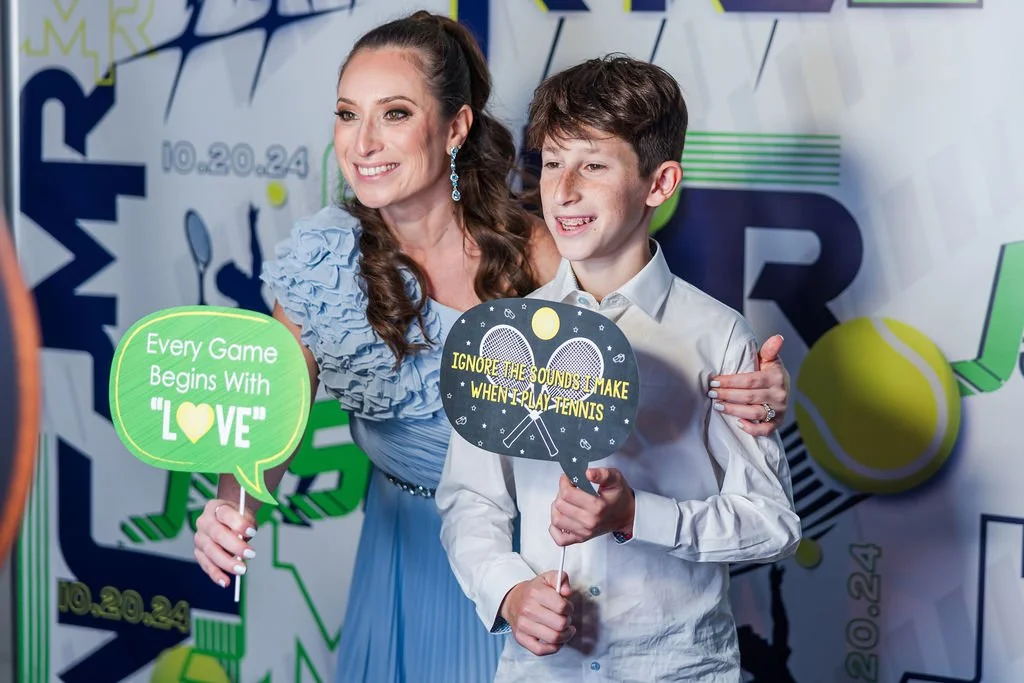 A woman and a boy holding signs at a tennis-themed event, smiling for a photo. The woman’s sign reads, "Every Game Begins With LOVE," and the boy’s sign says, "Ignore the sounds I make when I play tennis." Behind them is a colorful backdrop with tenn