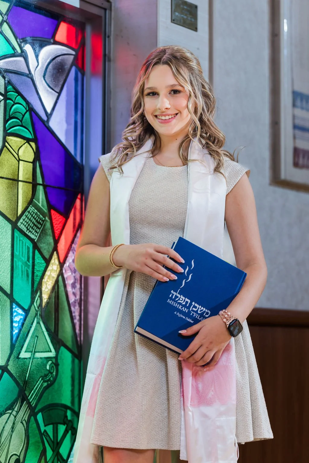 A young woman smiling and holding a blue book in front of a colorful stained glass window, dressed in a light-colored dress and a white vest in an indoor setting.